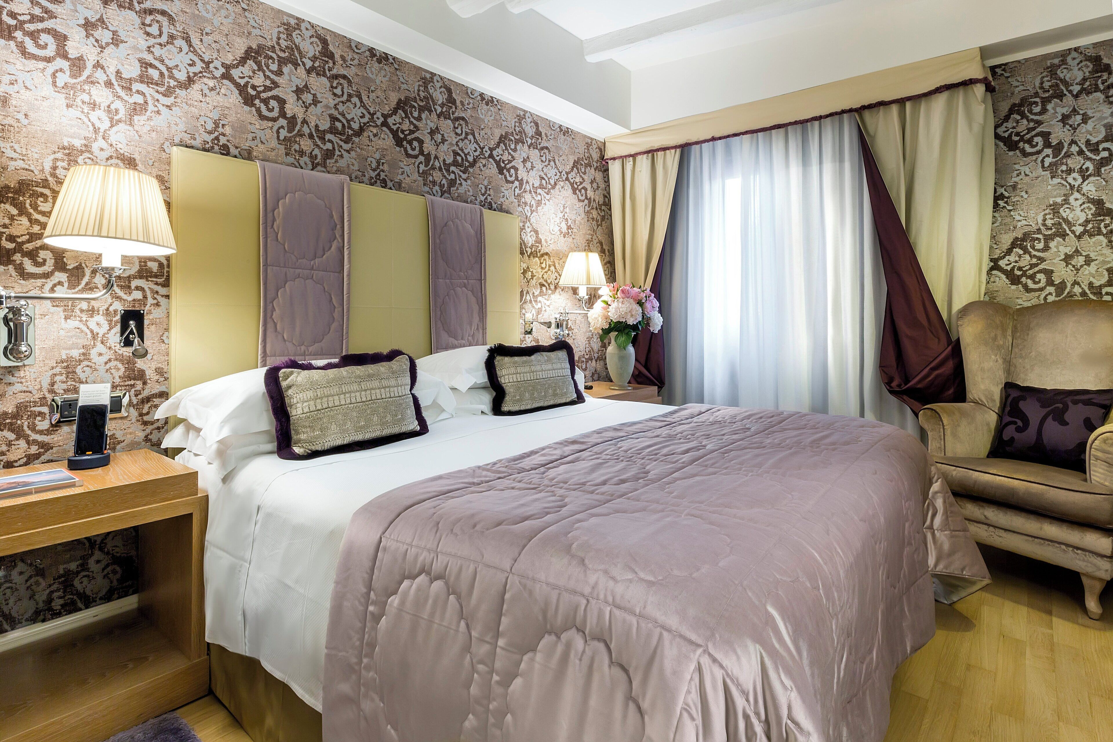 A hotel room with a large bed, a nightstand with a lamp, a chair, and a wall with patterned wallpaper.