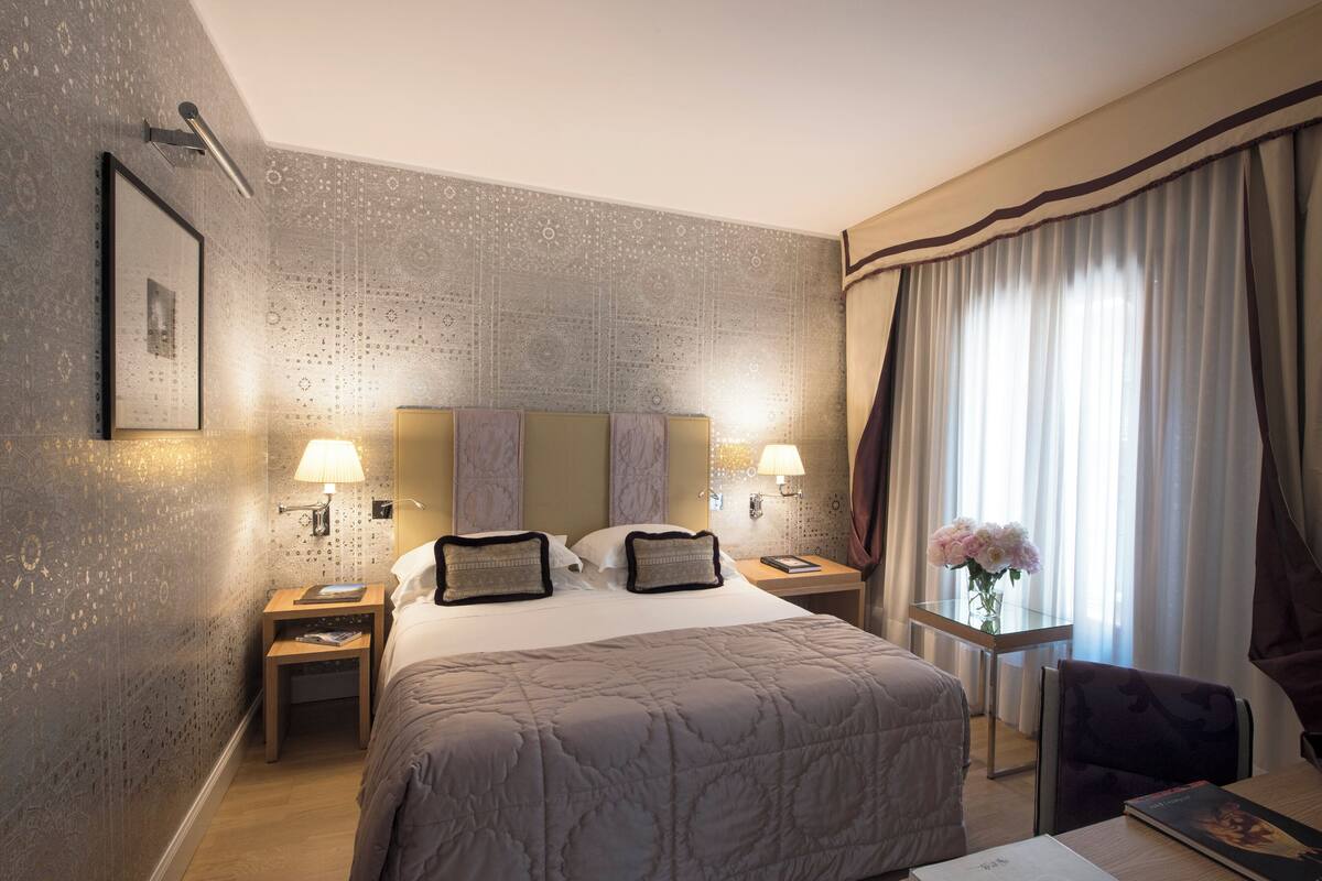 A hotel room with a large bed, two bedside tables with lamps, a small table with a vase of flowers, and a chair.