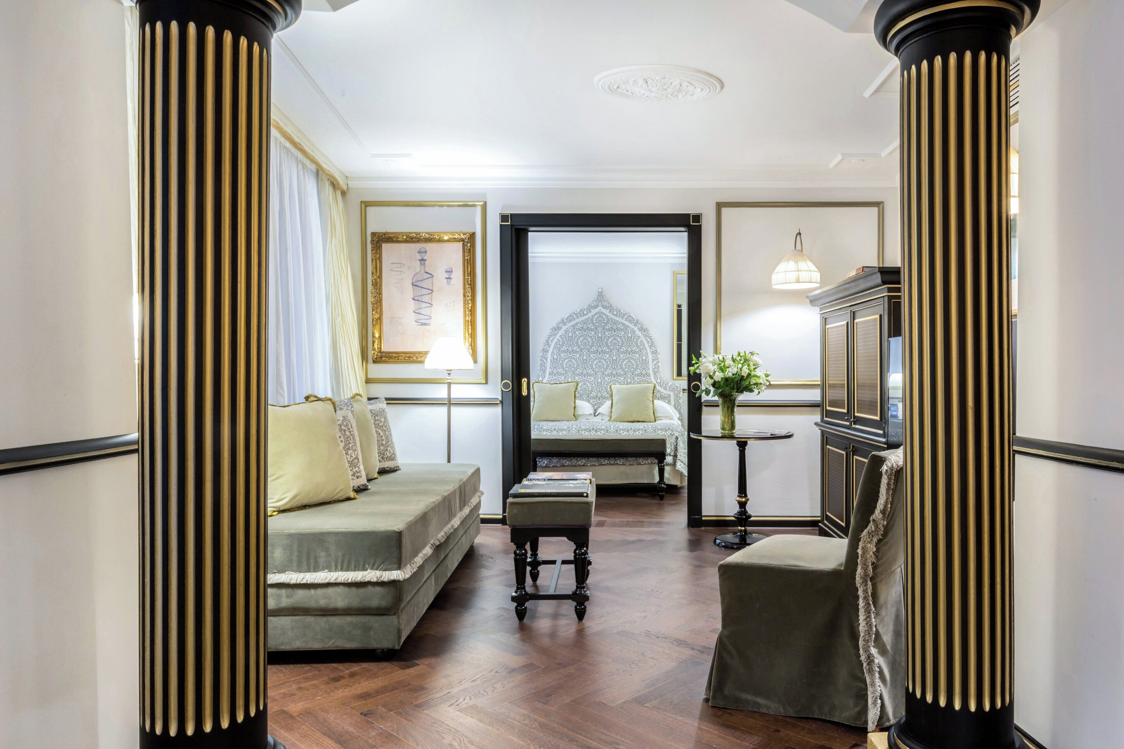 A hotel room with a bed, a sofa, a small table, and a chair. There is a large window with curtains, a lamp, and a framed picture on the wall.
