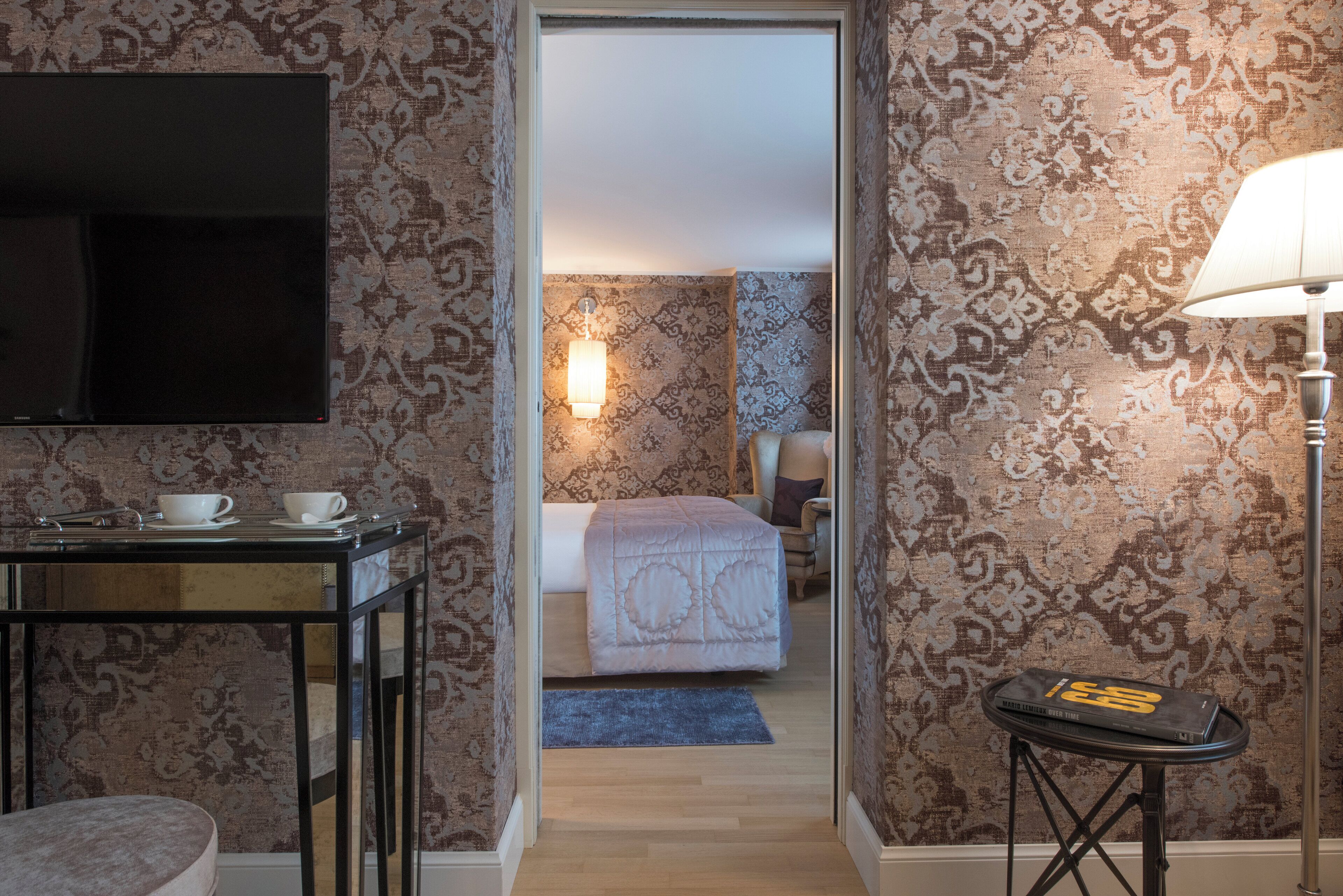 A hotel room with a bed, a desk with two cups, a lamp, and a TV mounted on the wall.