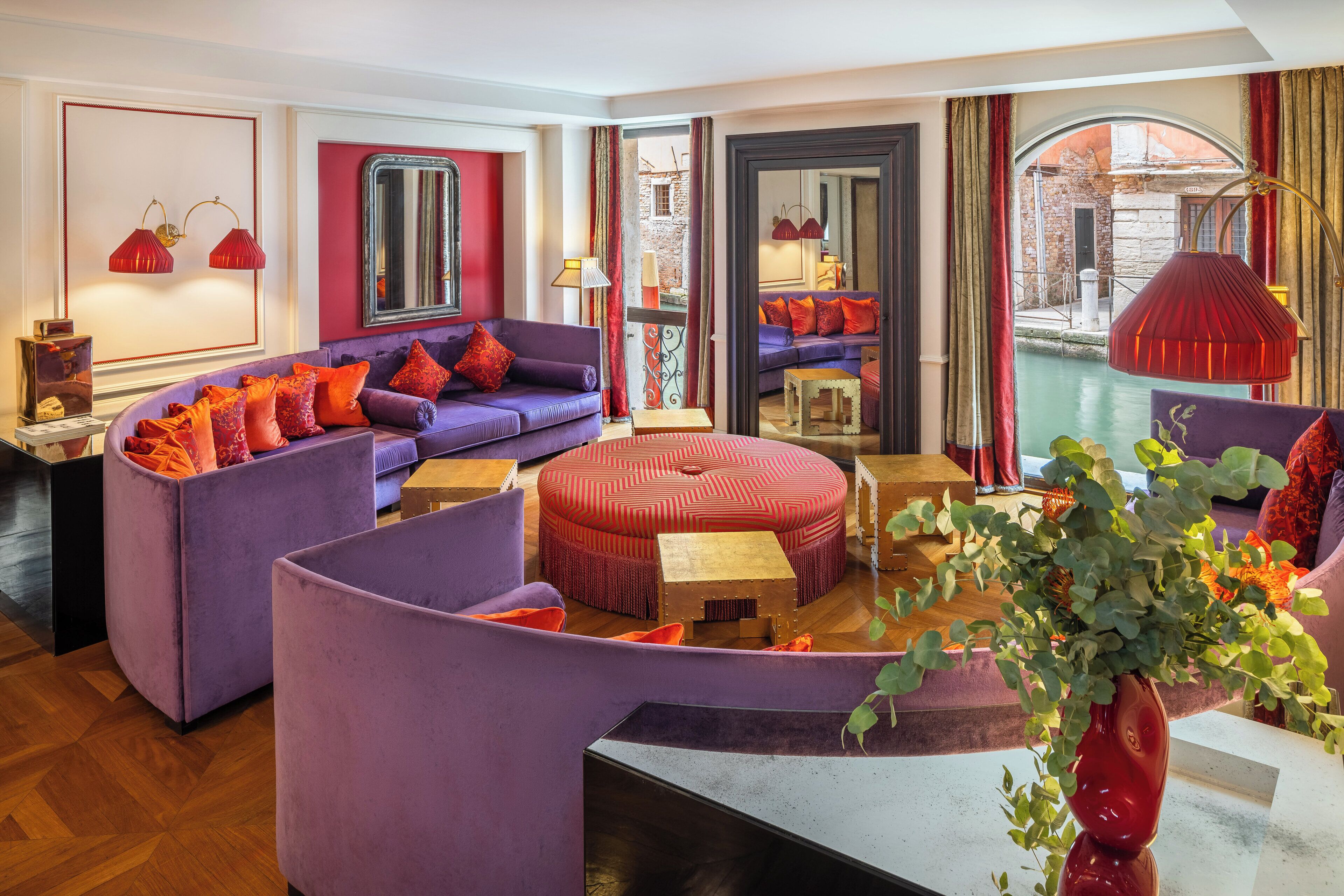 Un salon moderne avec des canapés violets, un pouf rouge et un vase rouge contenant des plantes vertes.