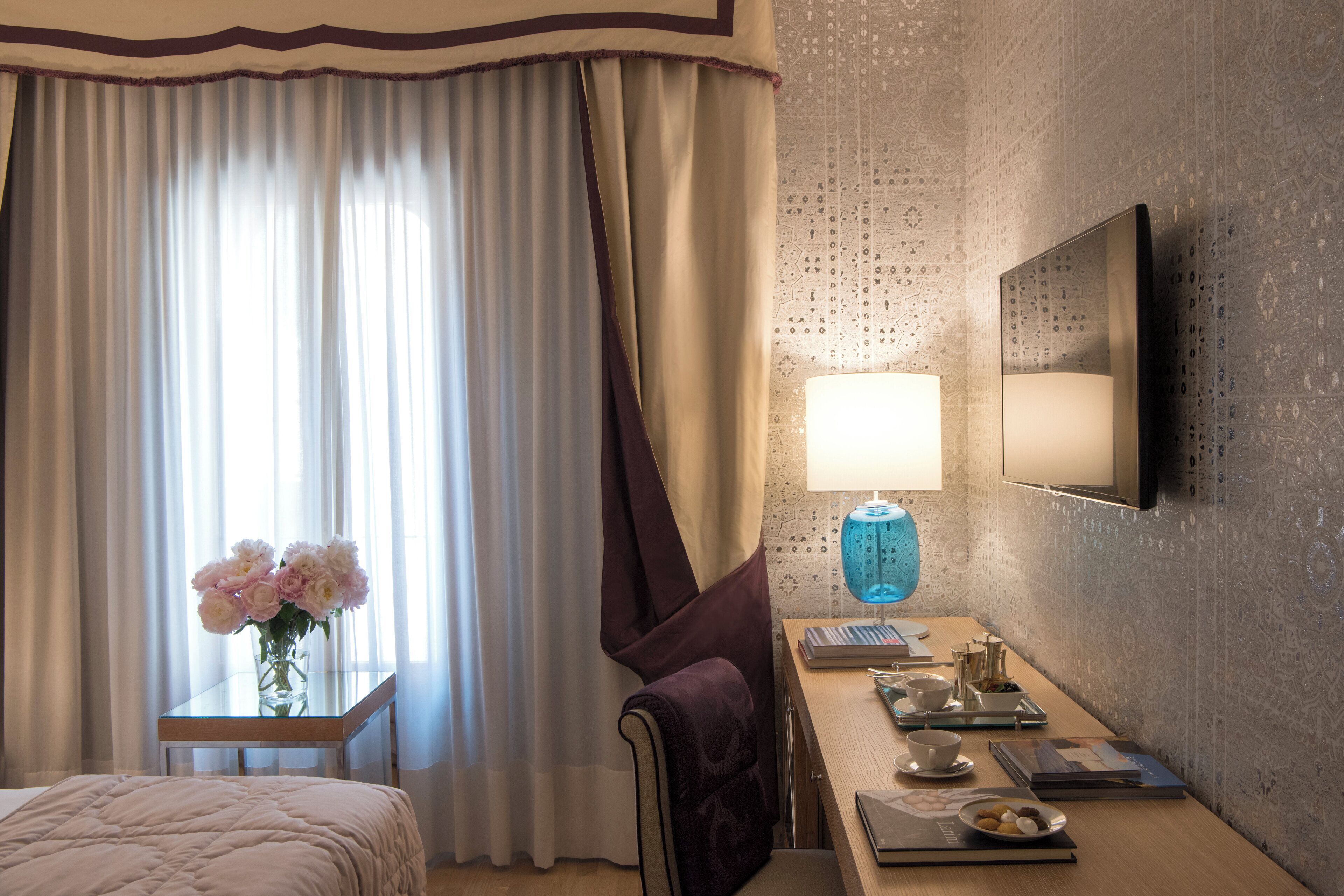 Habitación de hotel con cama, escritorio con lámpara, florero con flores y televisor montado en la pared.