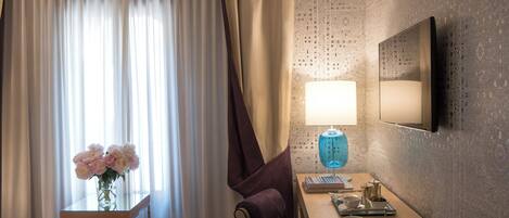 Habitación de hotel con cama, escritorio con lámpara, florero con flores y televisor montado en la pared.