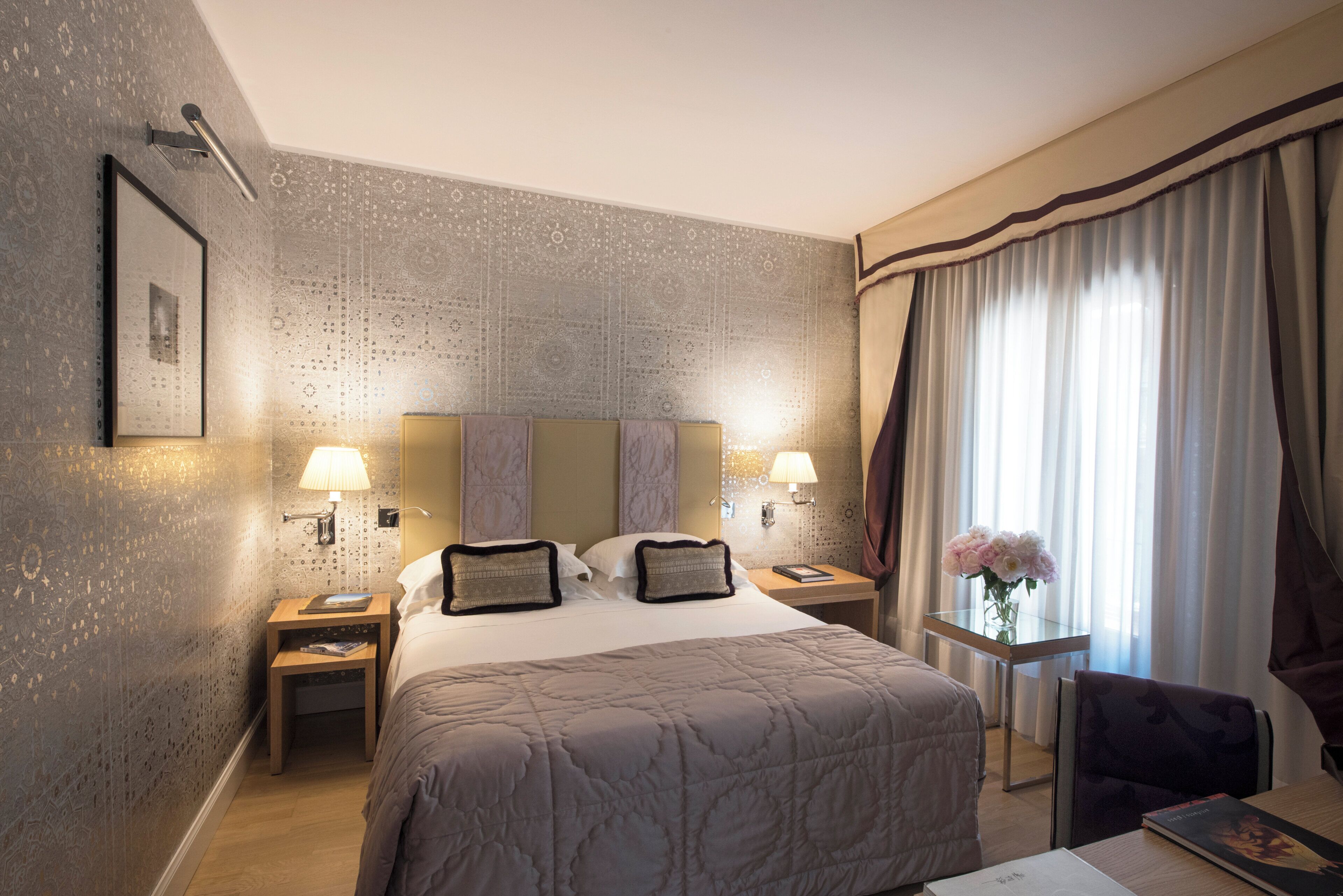 Une chambre d’hôtel avec un grand lit, deux tables de chevet avec des lampes, une petite table sur laquelle est posé un vase de fleurs et un fauteuil.