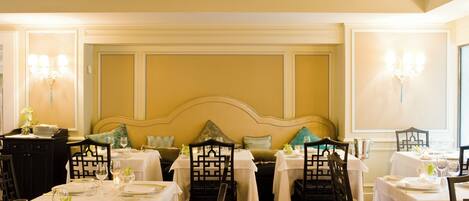 A dining area with tables set for service, featuring a high ceiling with wooden beams, a decorative wall panel, and a large cushioned headboard.