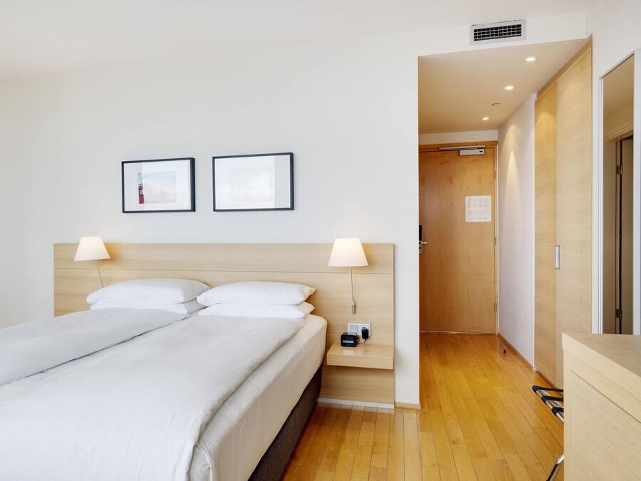 Una habitación de hotel con una cama, dos lámparas de noche, una mesita de noche y un cabecero de madera.