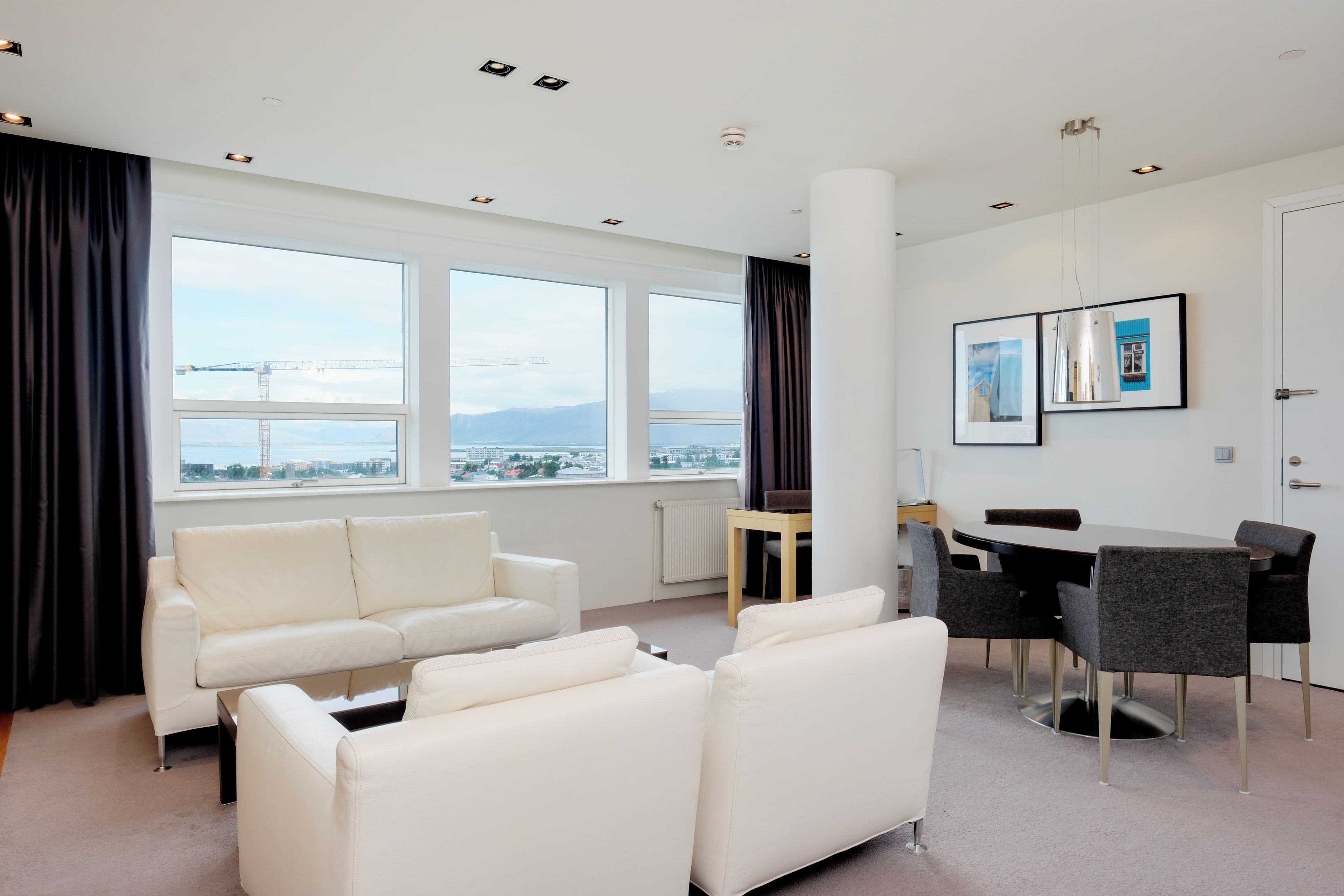 Quarto de hotel moderno com sofá, mesa de jantar, cadeiras e vista para a paisagem urbana.