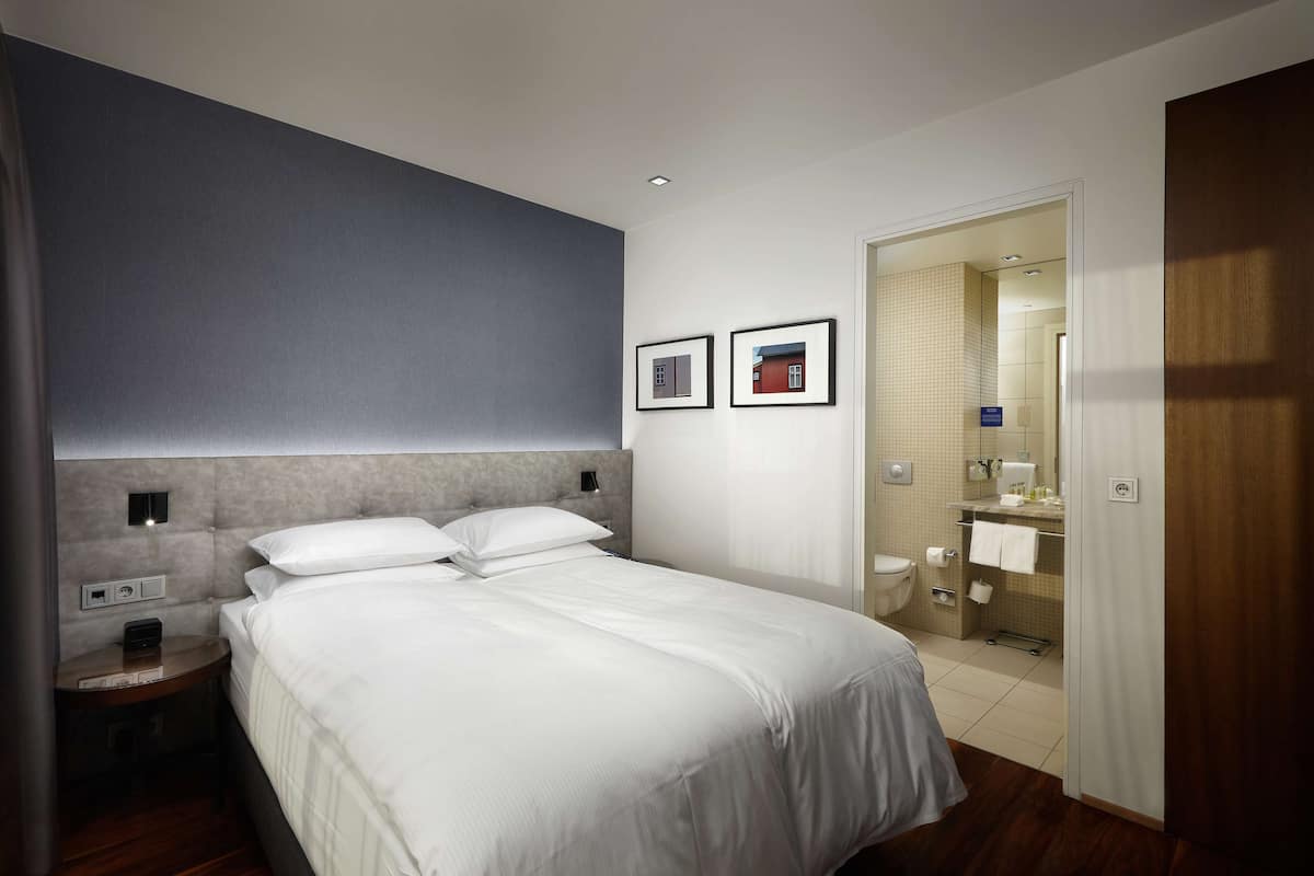 A modern hotel room with a large bed, a bedside table, a nightstand, a wall-mounted light, and a framed picture.
