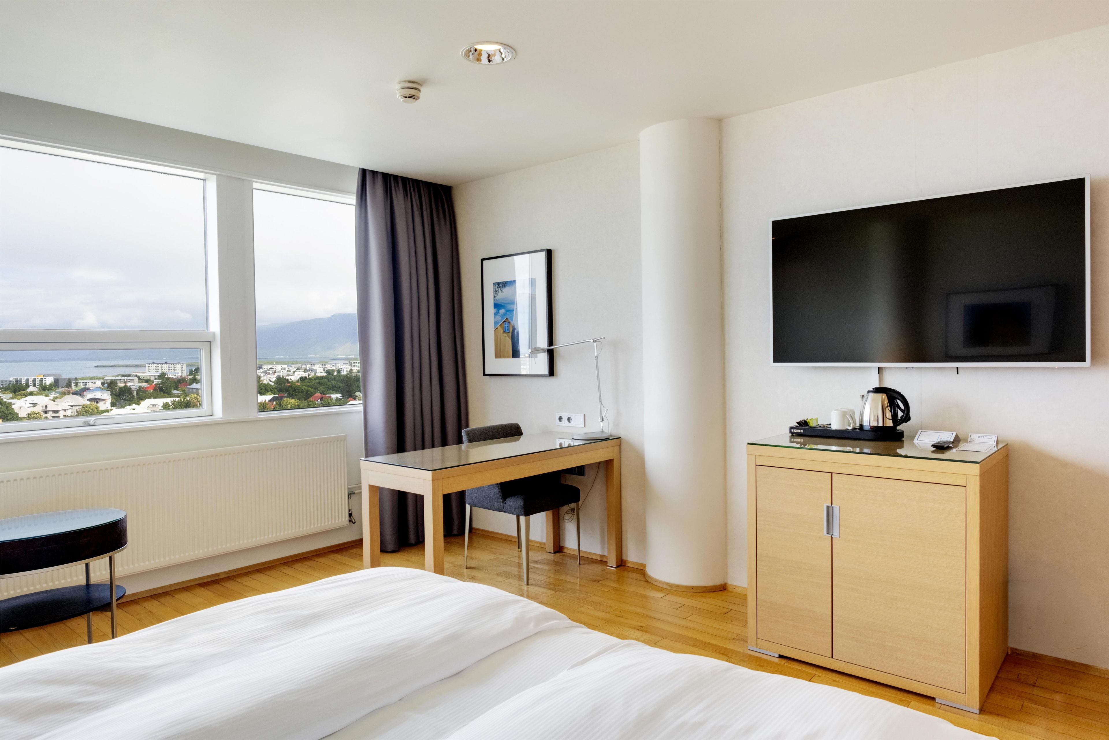 Quarto de hotel com cama, escrivaninha, cadeira, televisão e vista para a paisagem urbana.