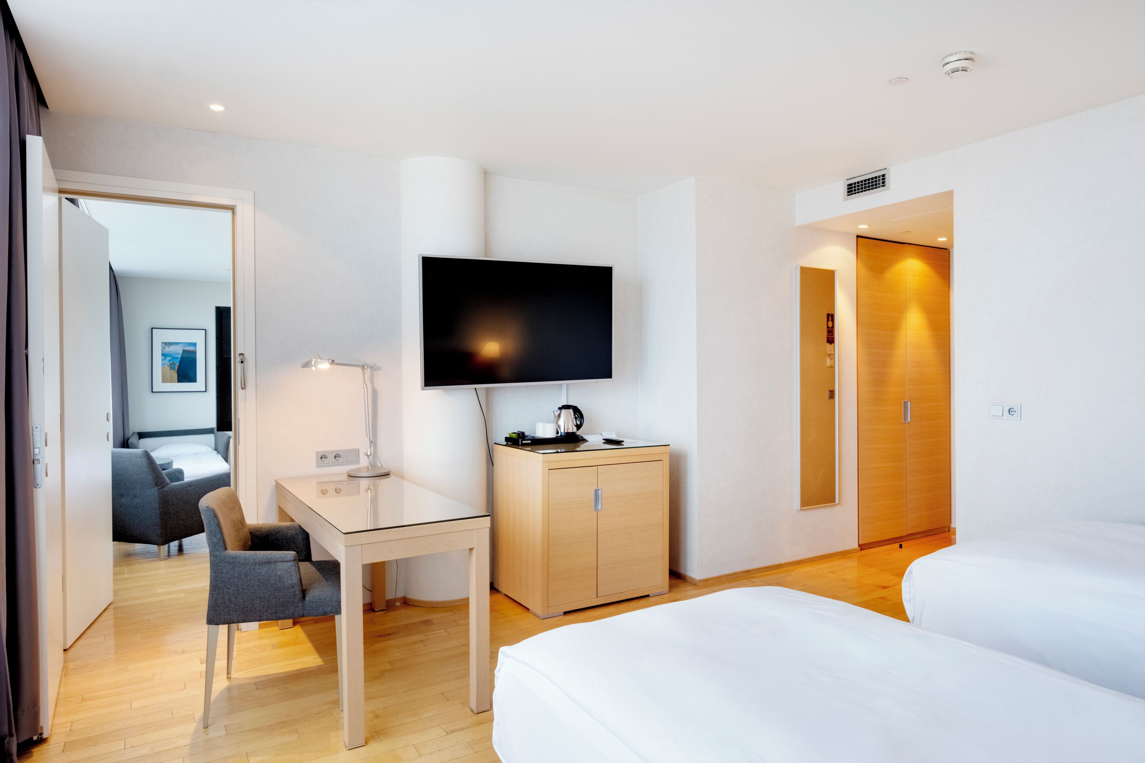 Ett hotellrum med en säng, ett skrivbord med en lampa, en tv, en garderob och en dörr som leder till ett annat rum.