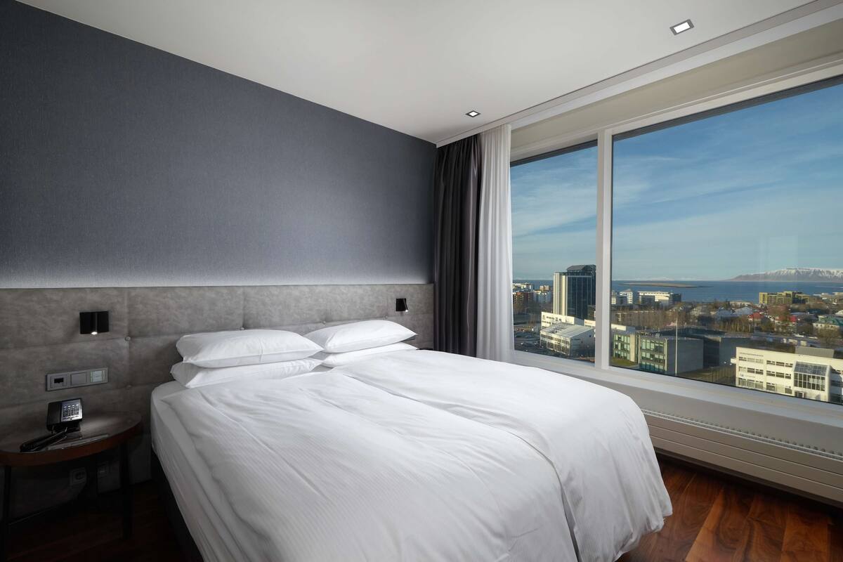 A modern hotel room with a large bed, a nightstand with a phone, and a city view through a large window.
