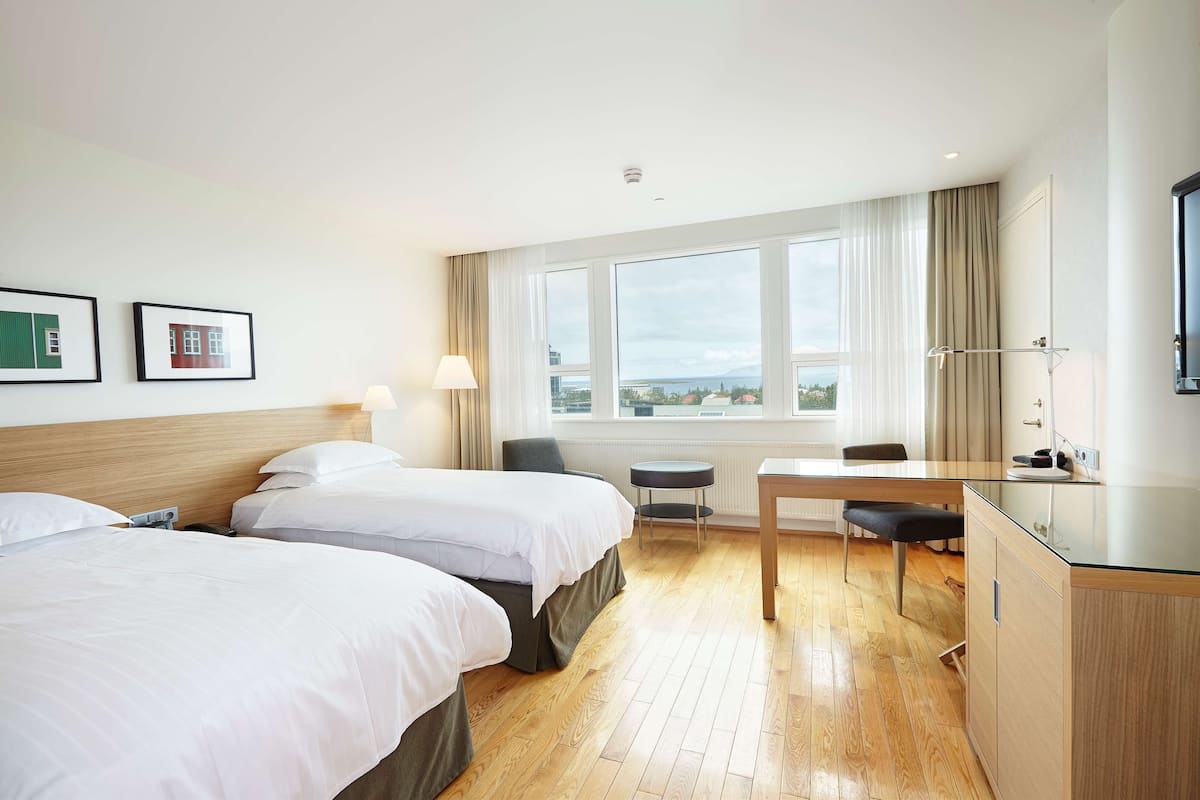 A hotel room with two beds, a desk, a chair, a small table, a TV, and a window with a view.