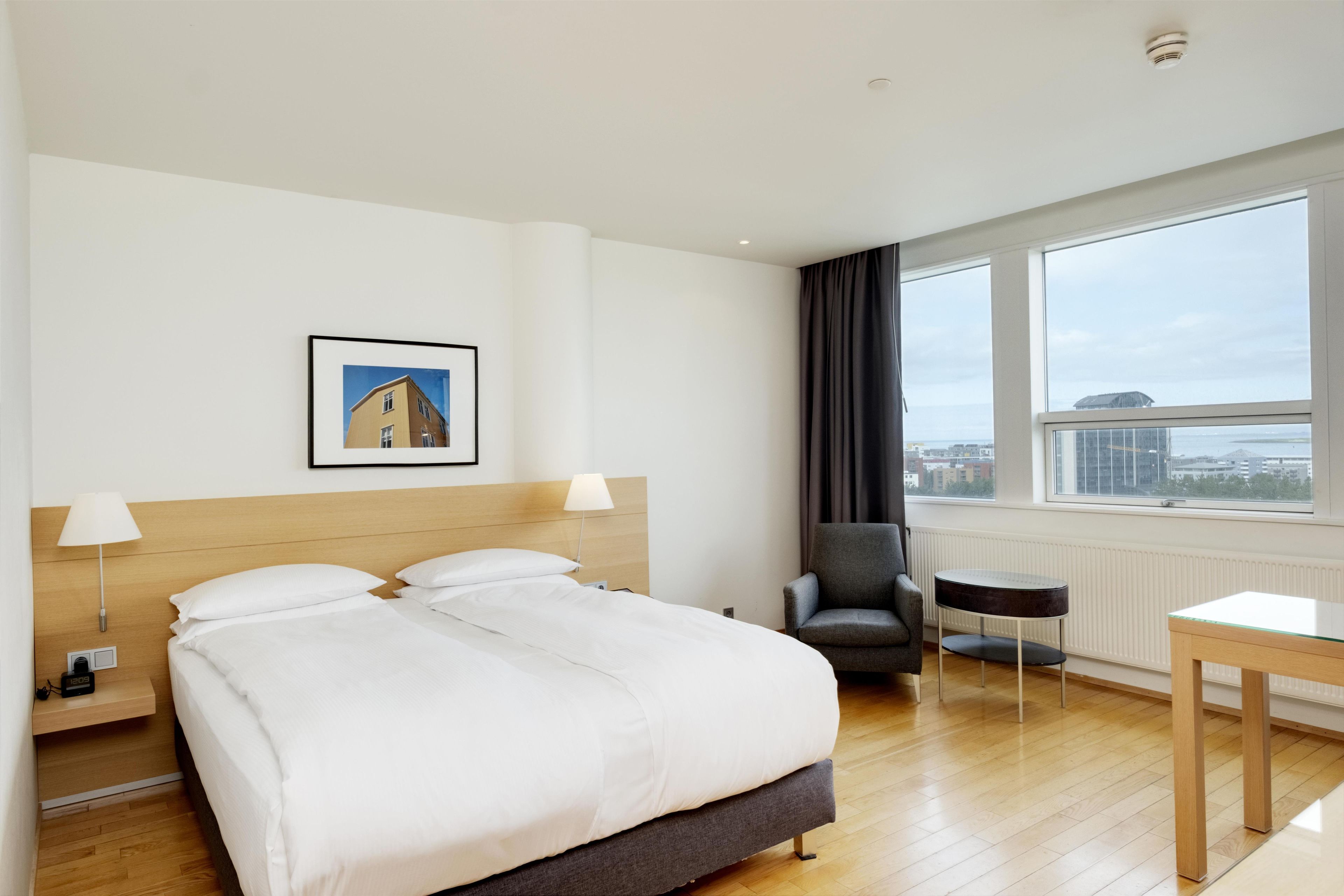 A hotel room with a large bed, a chair, a small table, a picture on the wall, and a window with a view.