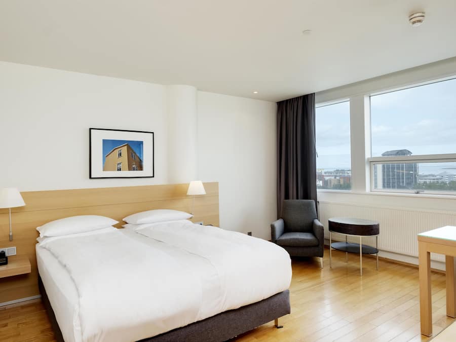 A hotel room with a large bed, a chair, a small table, a picture on the wall, and a window with a view.