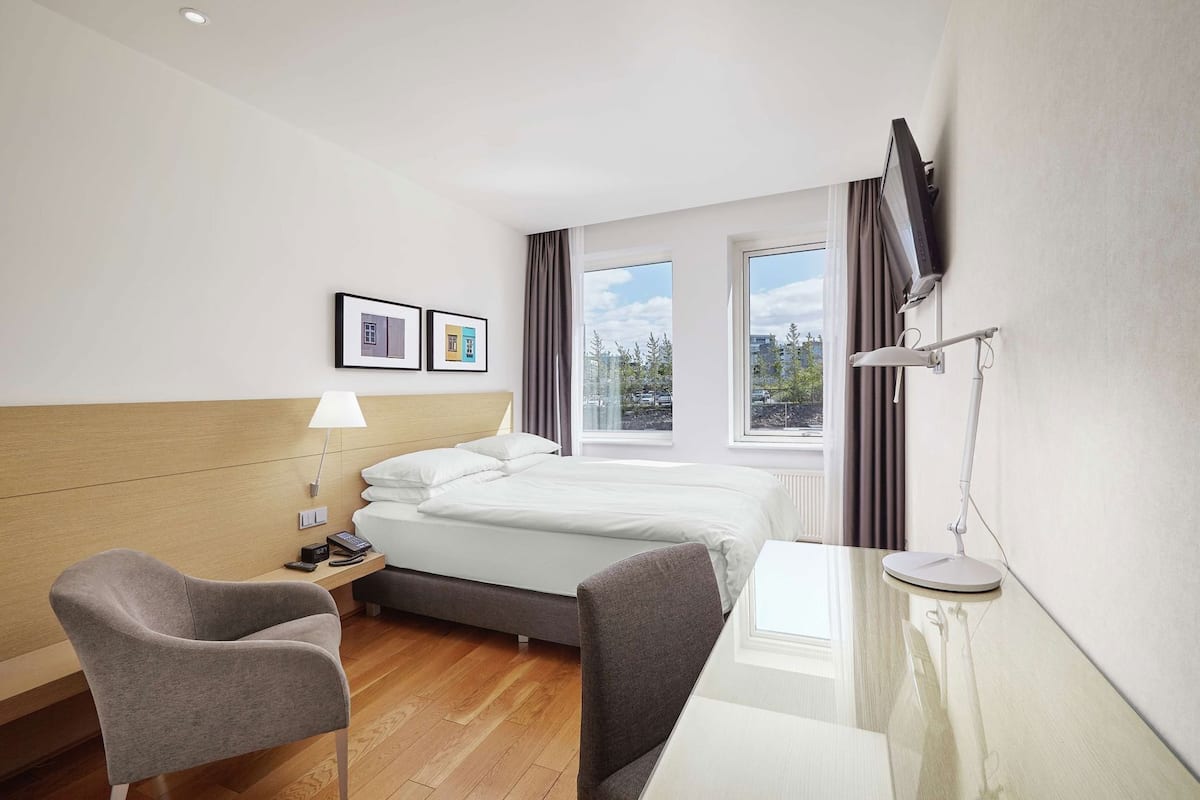 A hotel room with a bed, bedside table, lamp, desk, chair, and a TV mounted on the wall.