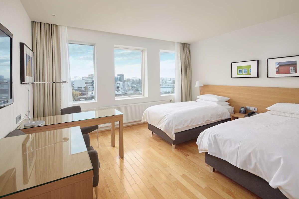 A hotel room with two beds, a glass desk, a chair, a nightstand, and large windows with a city view.