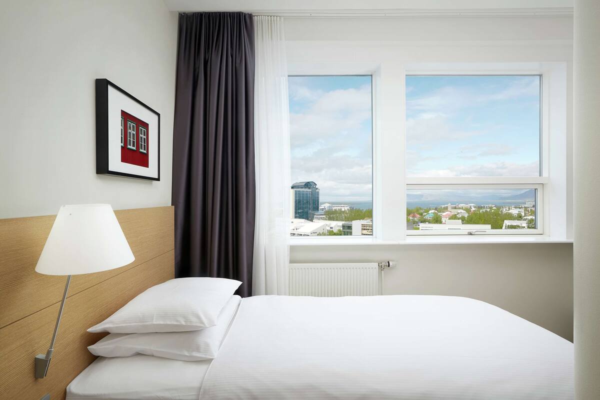 A hotel room with a bed, a nightstand with a lamp, a framed picture on the wall, and a window with a city view.