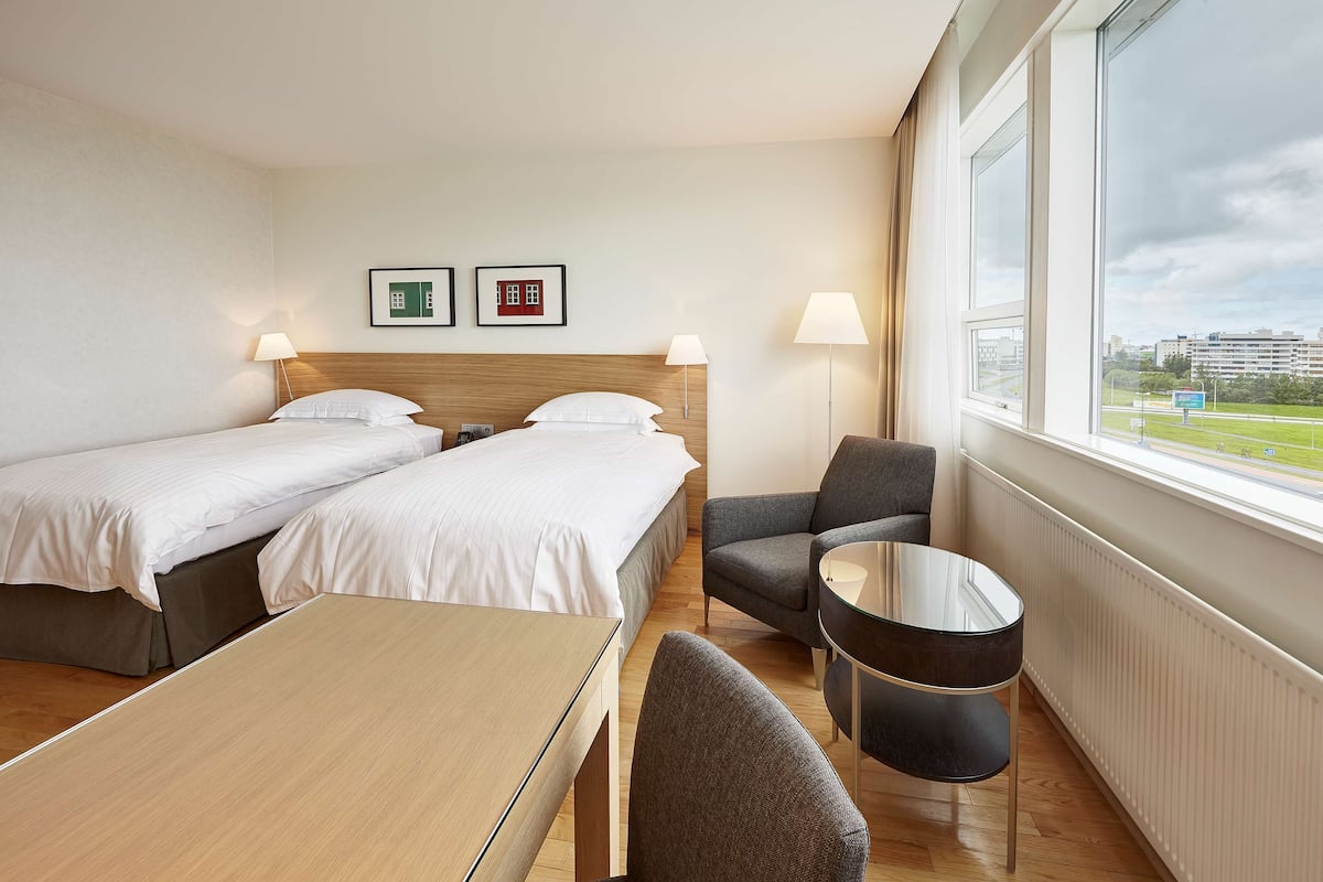 A hotel room with two beds, a small table, a chair, and a view of the city.