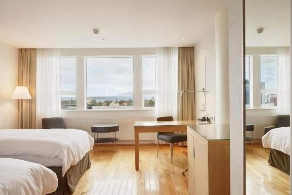 A hotel room with two beds, a desk, a chair, and a large window with curtains.