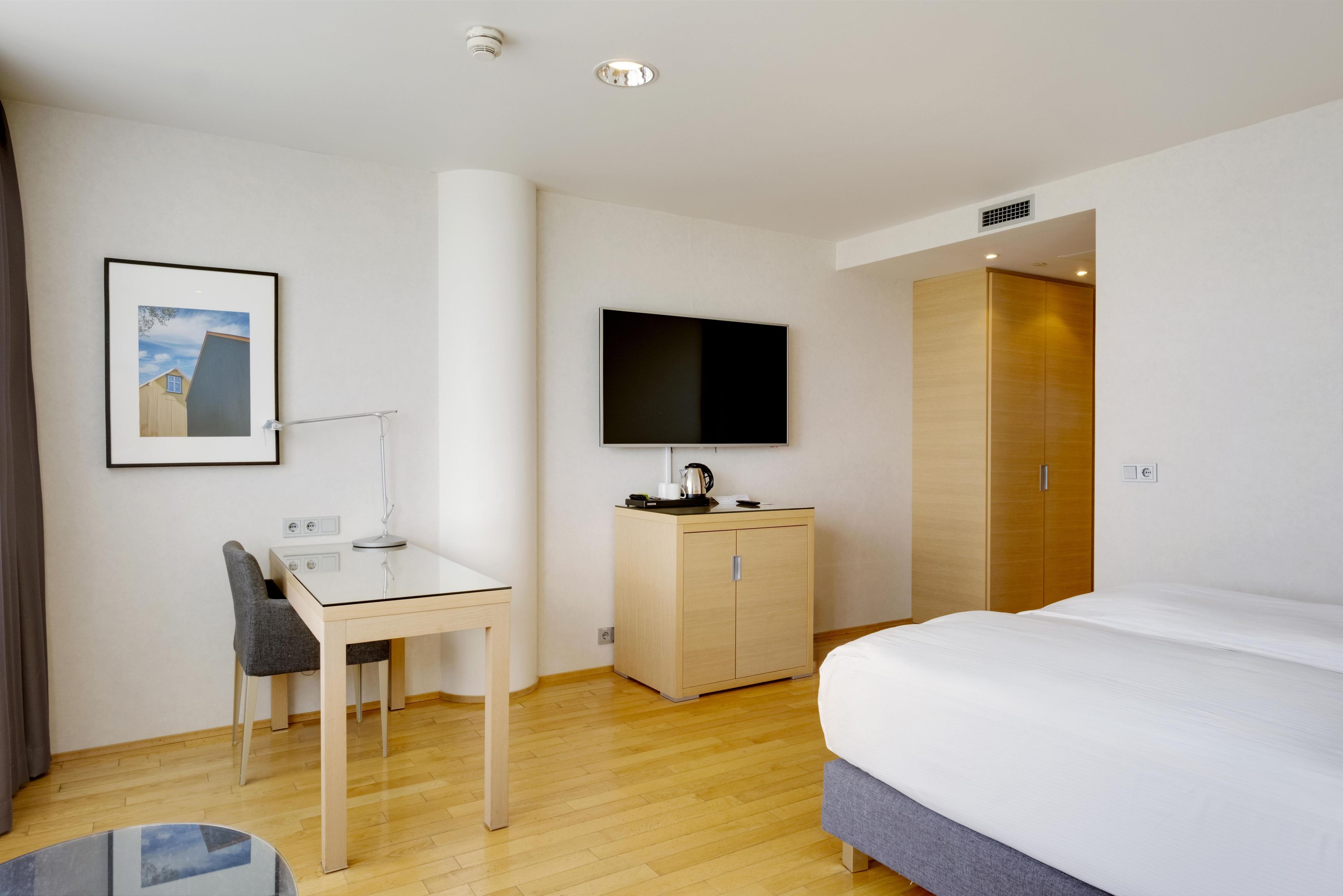 Quarto de hotel com uma cama, escrivaninha com cadeira, televisão, guarda-roupa e uma moldura na parede.