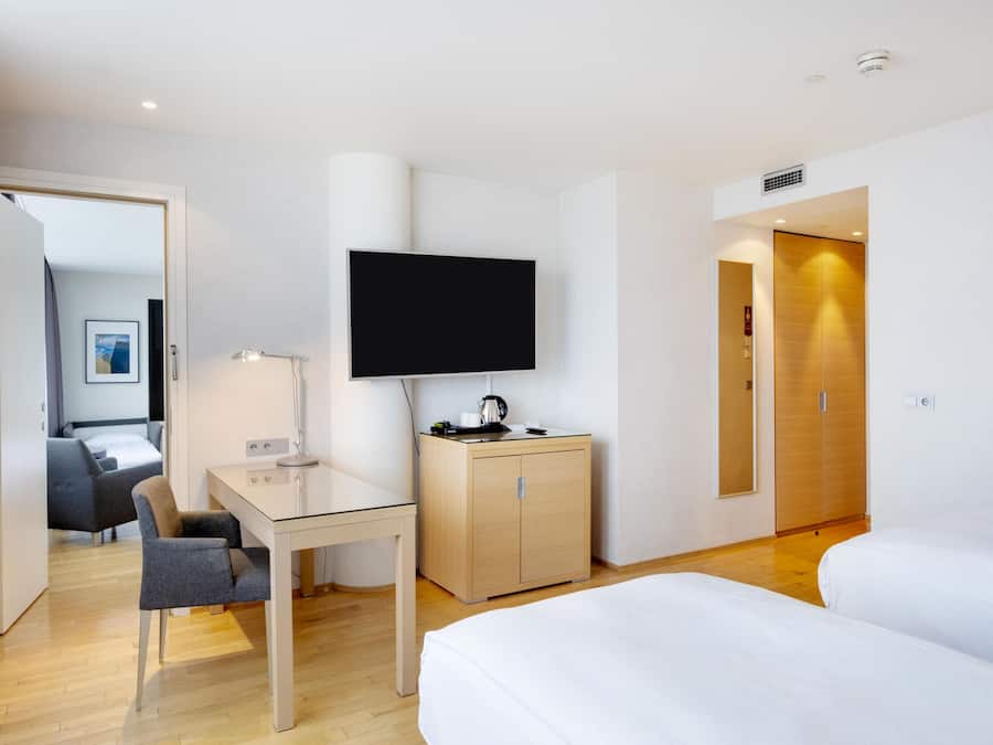 A hotel room with a bed, a desk with a chair, a television, a wardrobe, and a door leading to another room.