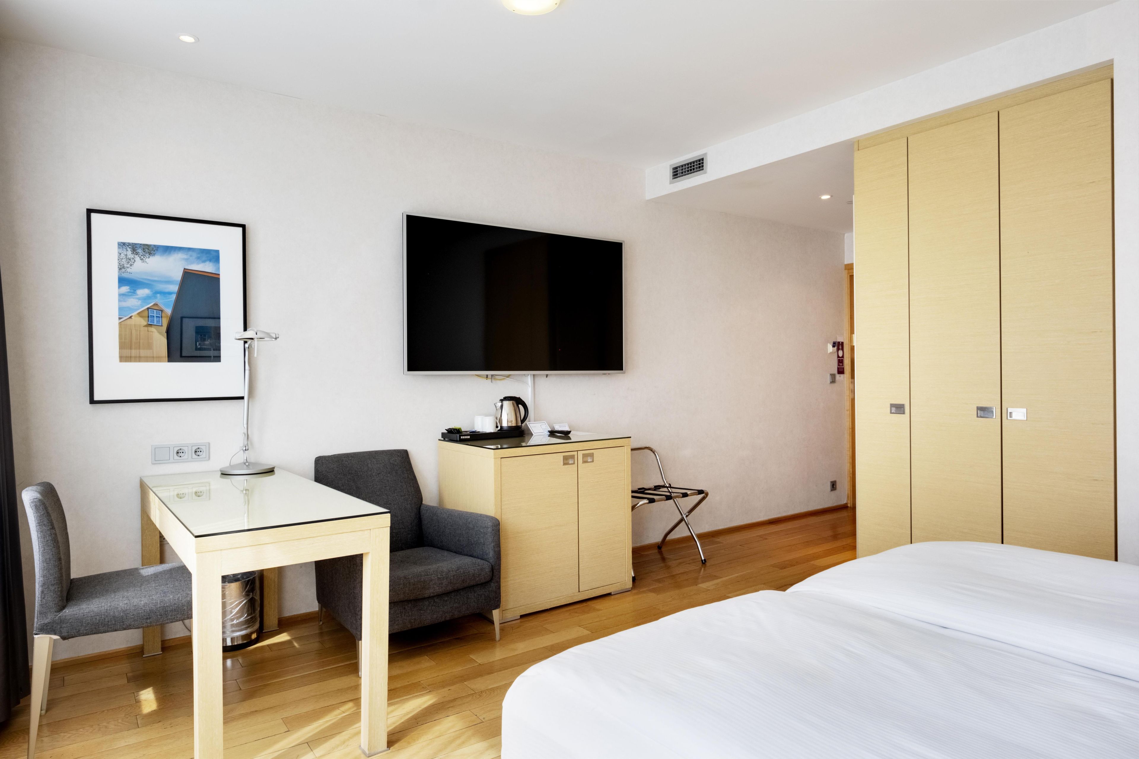 A hotel room with a bed, a desk with a chair, a television, a wardrobe, and a framed picture on the wall.