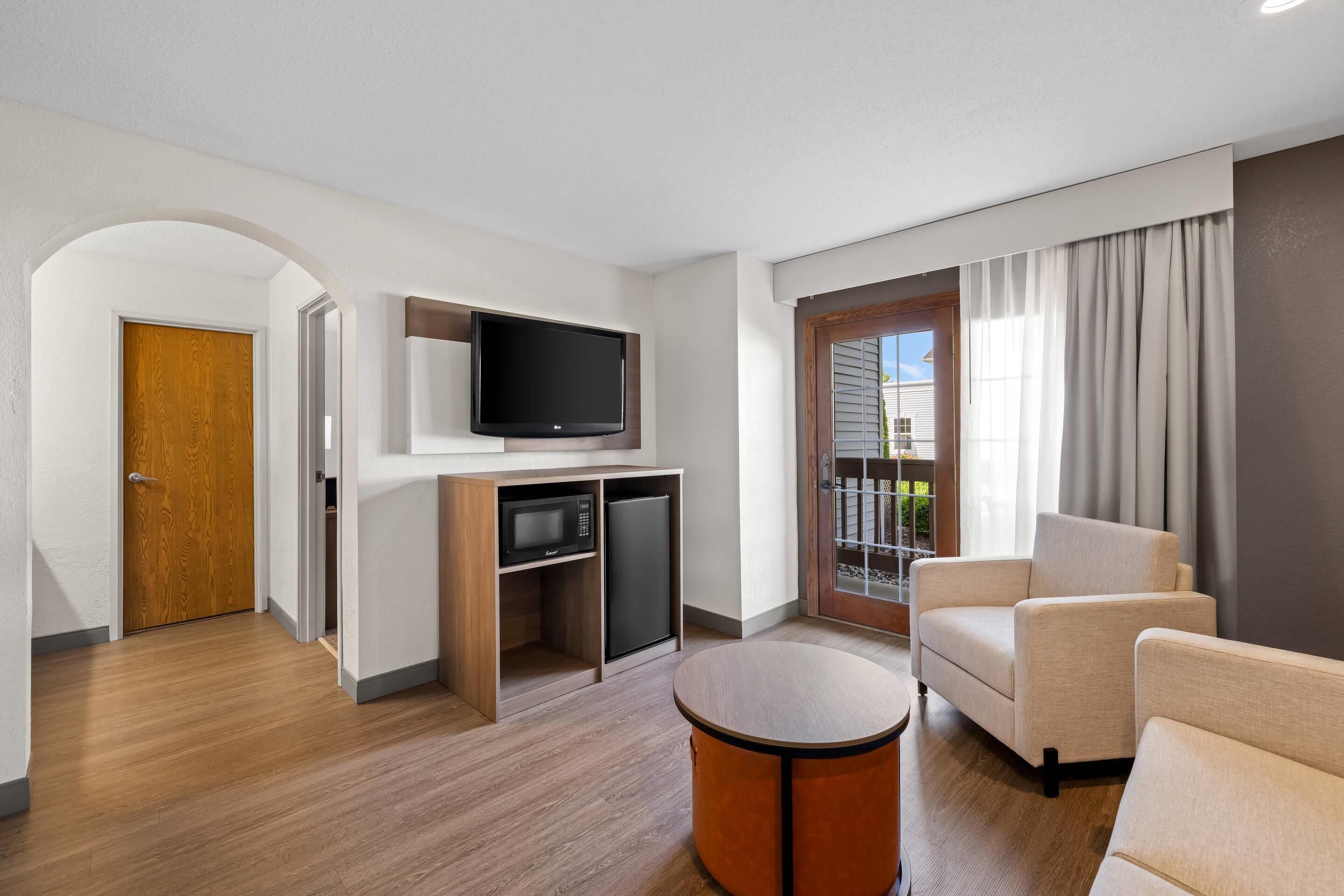 A modern living room with a flat-screen TV, a built-in microwave, a refrigerator, a round coffee table, and a beige sofa.