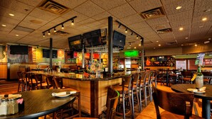 Een restaurantinterieur met meerdere eettafels en stoelen, een bar met een counter, en een televisie die aan het plafond is bevestigd.