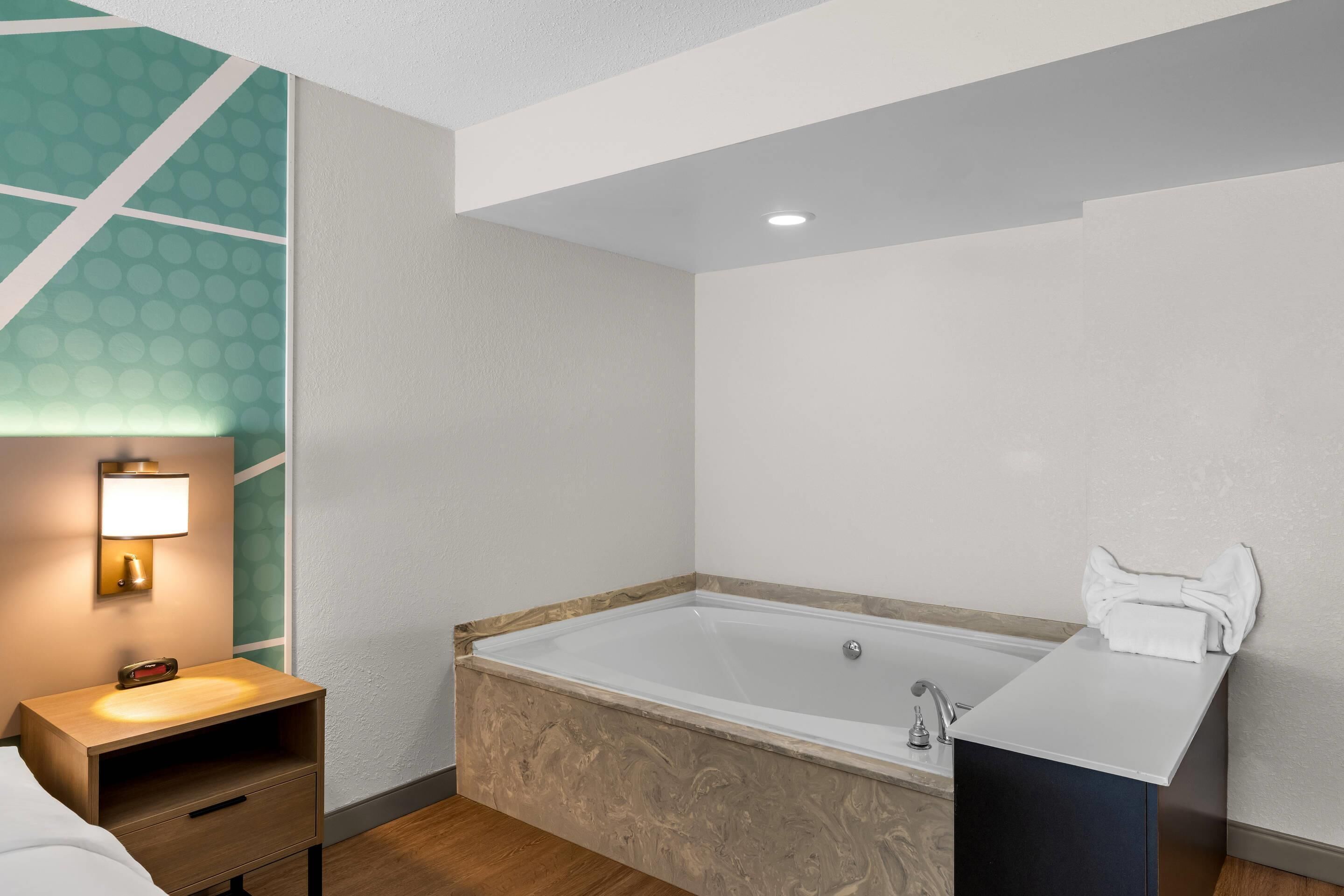 A modern bathroom with a bathtub, a vanity with a mirror, and a nightstand with a lamp.
