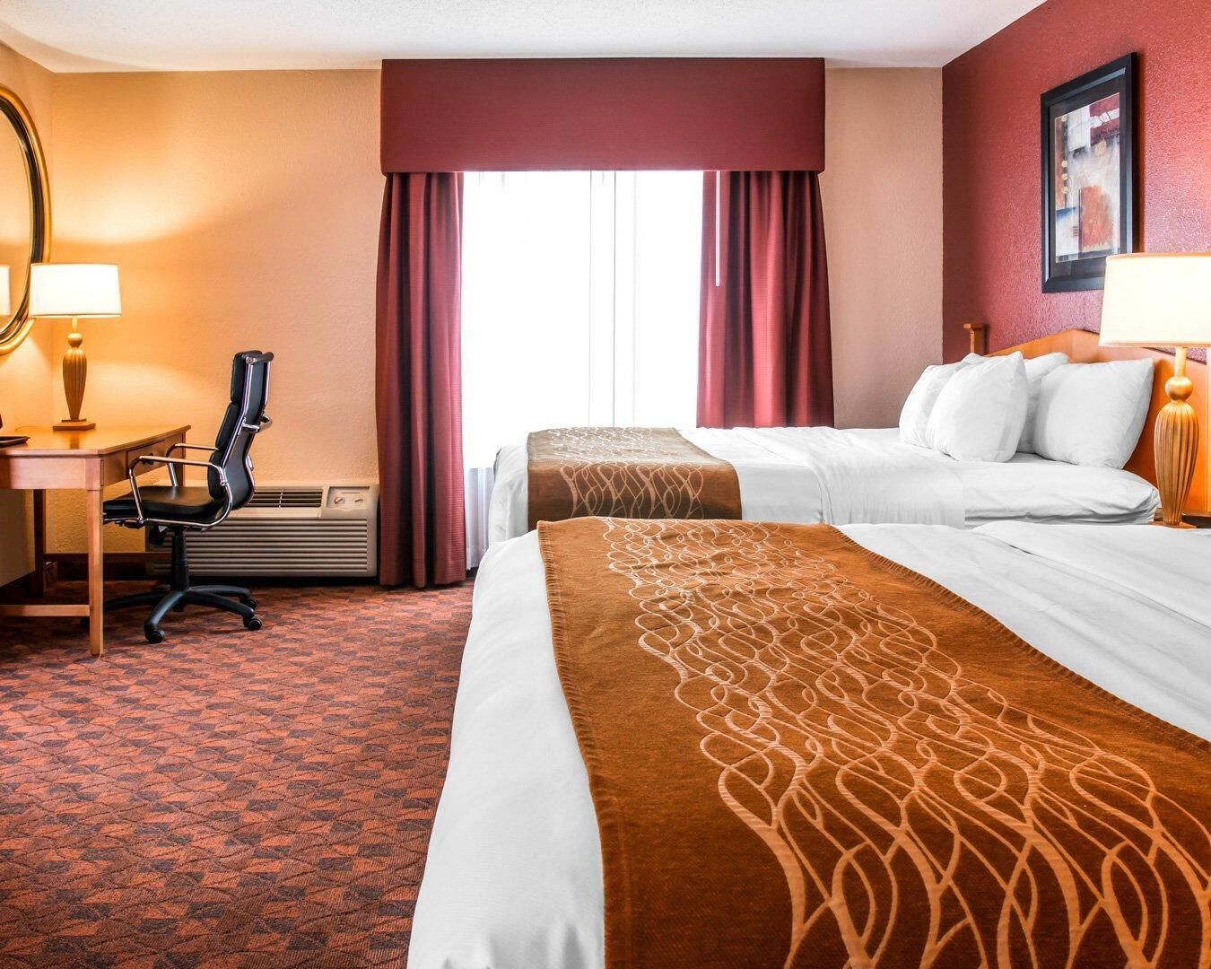 A hotel room with two beds, a desk with a chair, a lamp, and a large window with curtains.