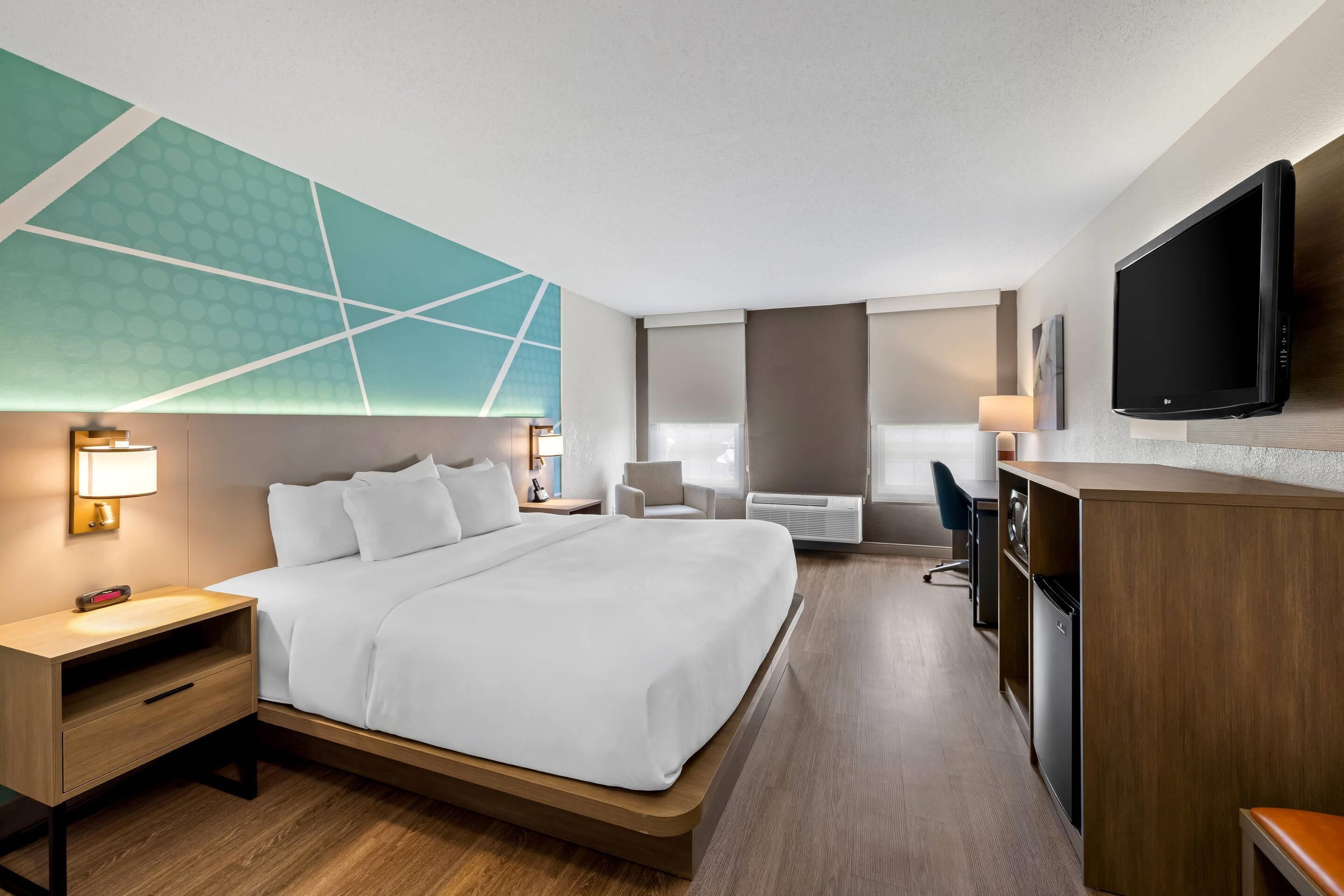 A hotel room with a bed, bedside table, TV, desk, chair, and a large window.