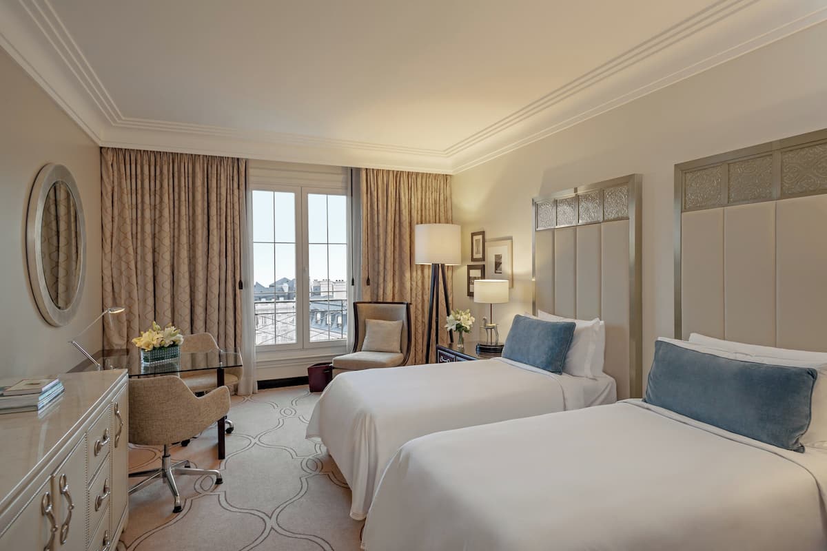 Habitación de hotel con dos camas, un escritorio, una silla y un gran ventanal con cortinas.