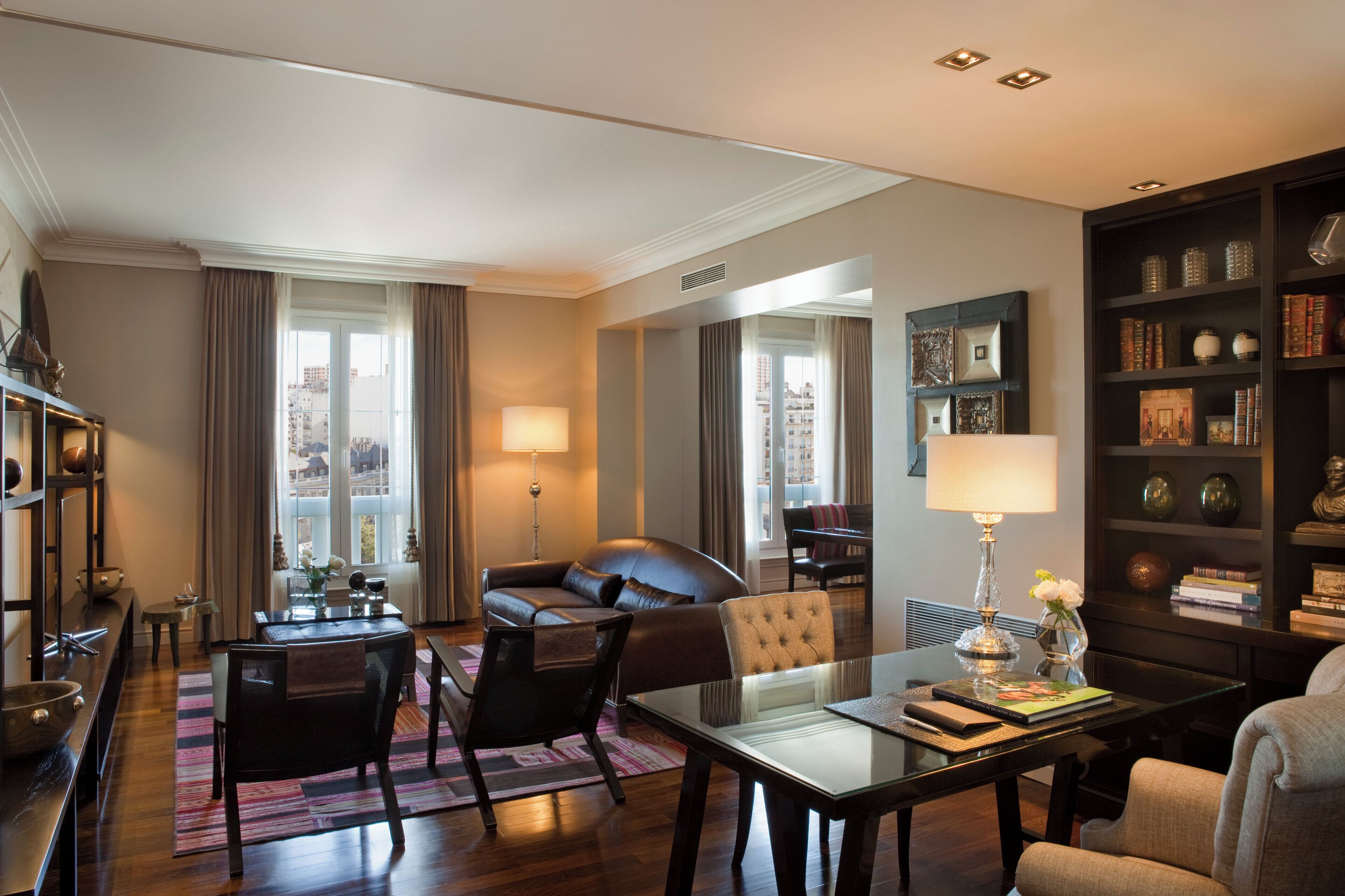 Una habitación de hotel moderna con zona de comedor, sofá, estantería y un ventanal con cortinas.