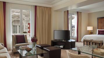 Quarto de hotel com mesa de centro de vidro, sofá, televisão, cama e vista para a cidade.