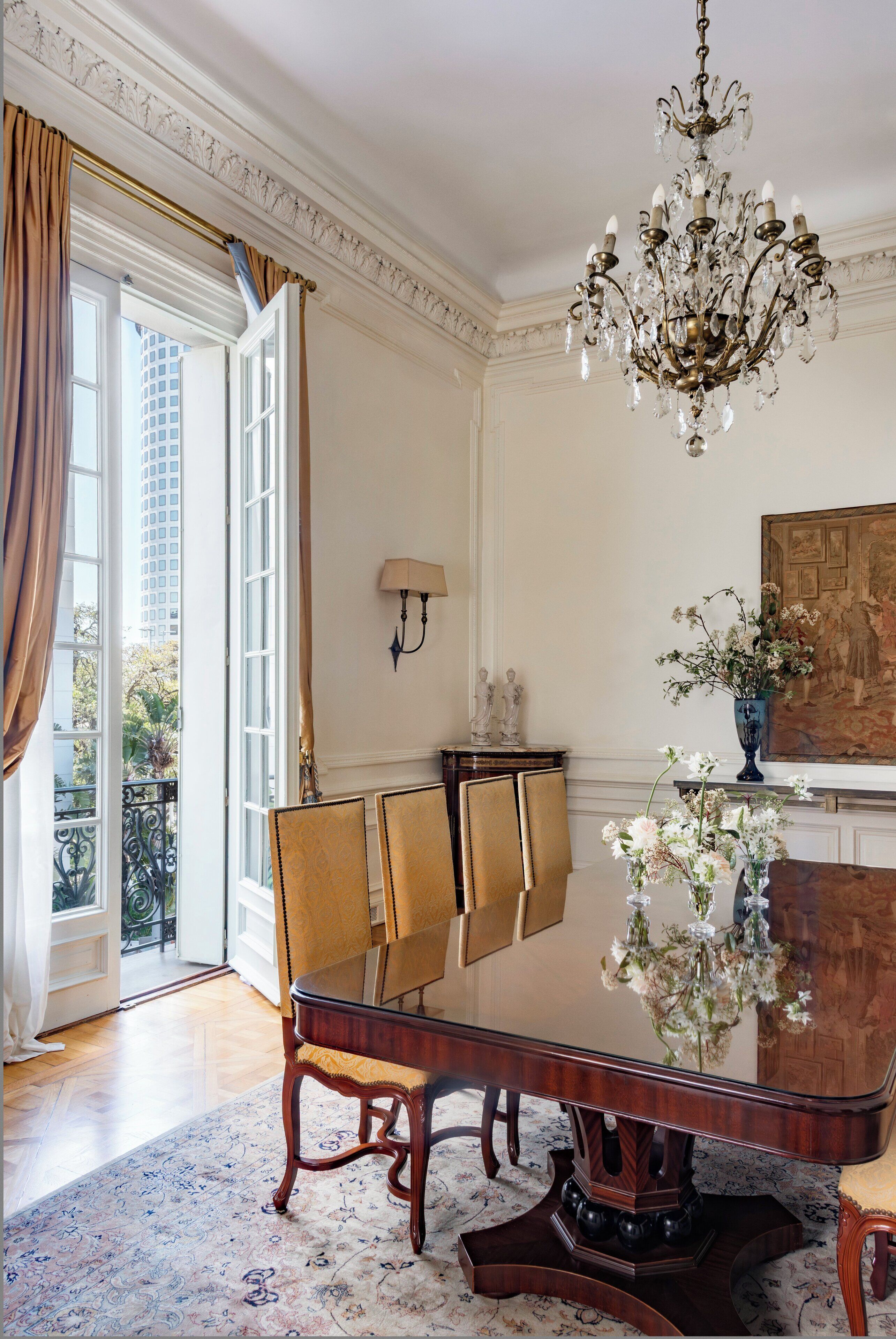 Uma sala de jantar com uma grande mesa de madeira, cadeiras, um candelabro e vista para o exterior por uma porta aberta.
