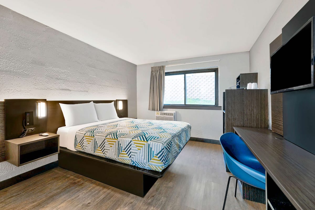 Ein modernes Hotelzimmer mit einem großen Bett, einem Schreibtisch, einem blauen Sessel, einem Fernseher und einem Fenster mit Vorhängen.