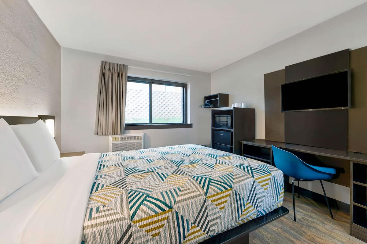 Ein Hotelzimmer mit einem großen Bett, einem Schreibtisch mit Stuhl, einem an der Wand montierten Fernseher und einem Fenster mit Vorhängen.