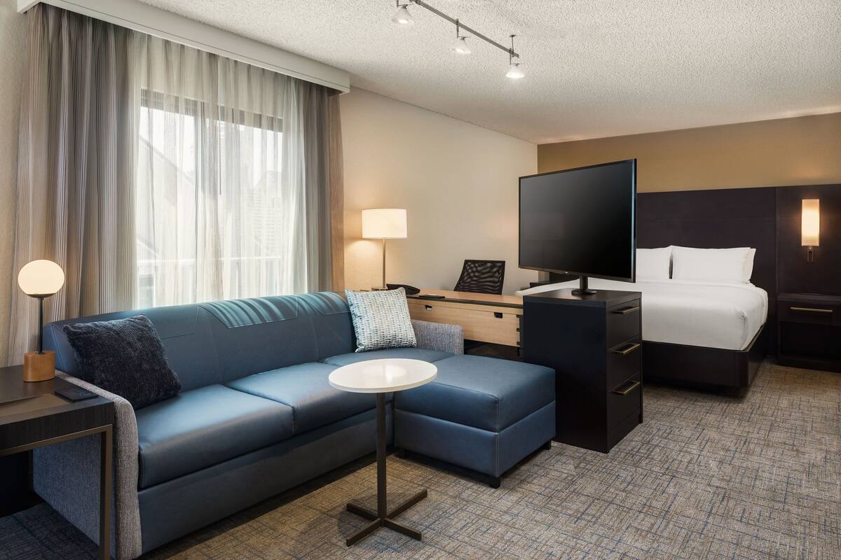 A hotel room with a blue sofa, a small round table, a bed, a desk with a TV, and a nightstand.