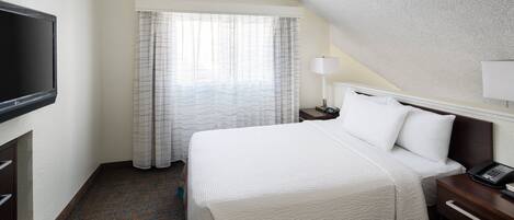 Habitación de hotel con cama, televisor, mesita de noche con lámpara y ventana con cortinas.
