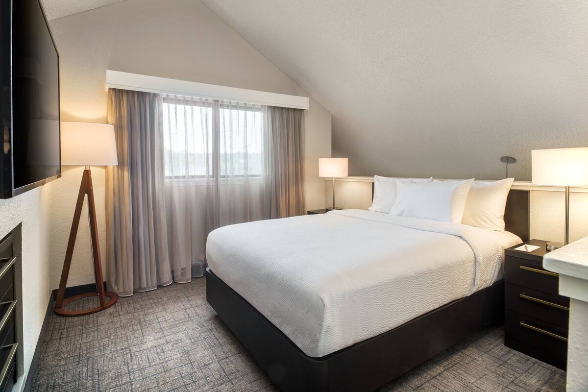 A hotel room with a large bed, a flat-screen TV, a bedside table with a lamp, and a window with curtains.