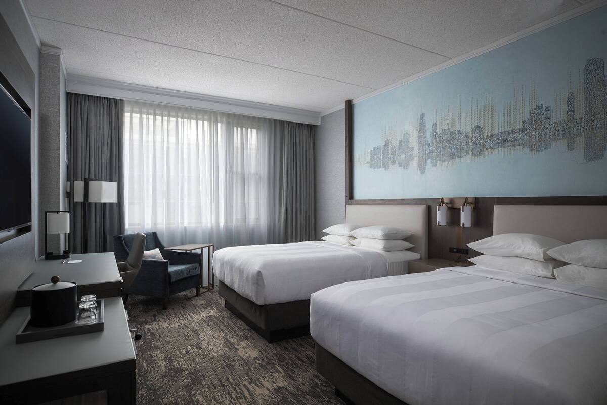 A hotel room with two beds, a desk, a chair, a TV, and a cityscape mural on the wall.