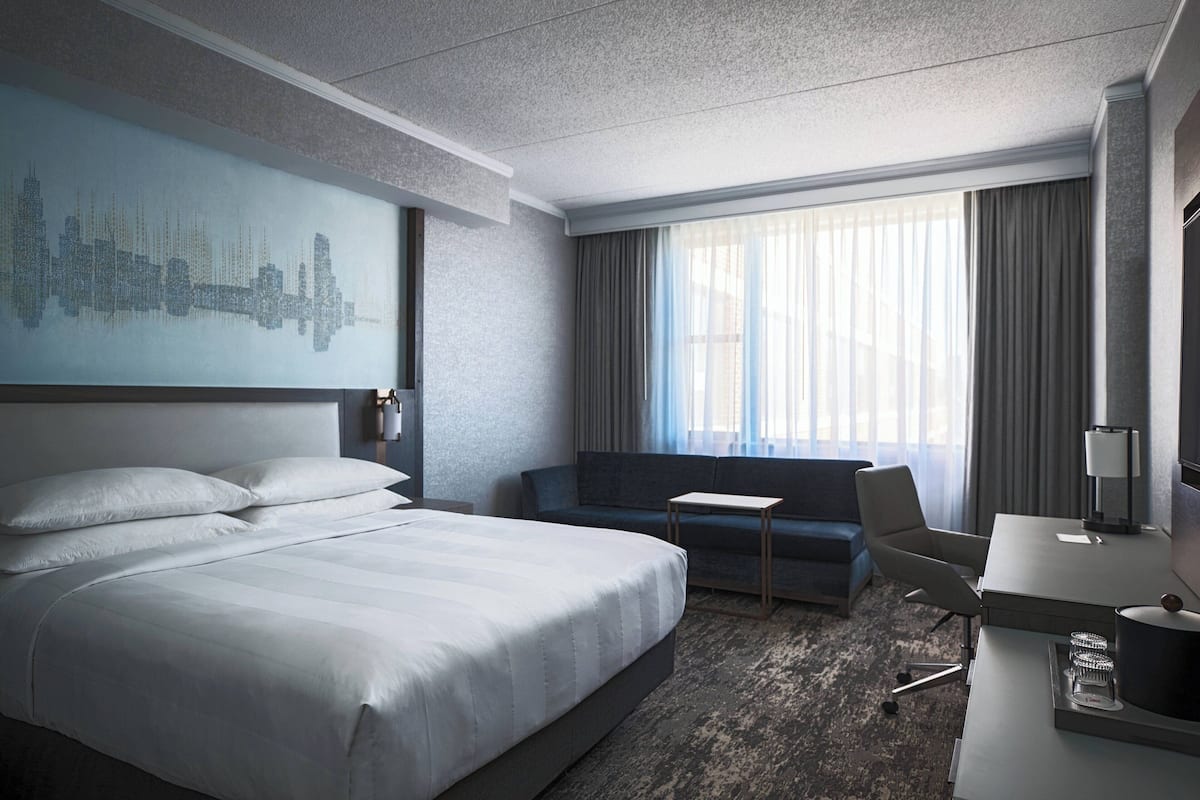 A modern hotel room with a large bed, a seating area, a desk, and a cityscape artwork on the wall.
