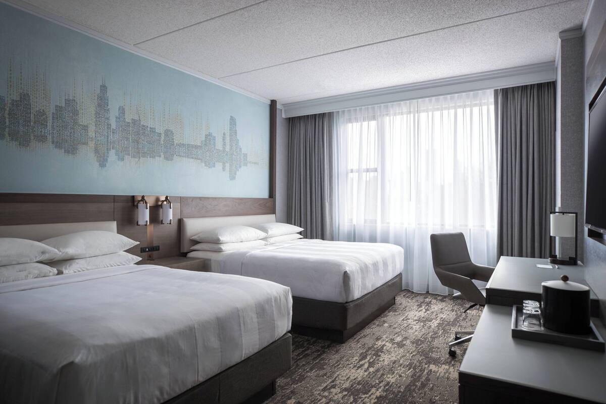 A hotel room with two beds, a desk, a chair, a cityscape mural, and a large window with curtains.