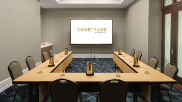 Une salle de conférence équipée d’une longue table, de chaises, d’un écran de projection affichant le logo « COURTYARD BY MARRIOTT », et d’un petit coin cuisine.
