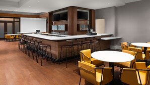 Uma área de bar moderna com um balcão central, vários bancos e mesas redondas com cadeiras amarelas.