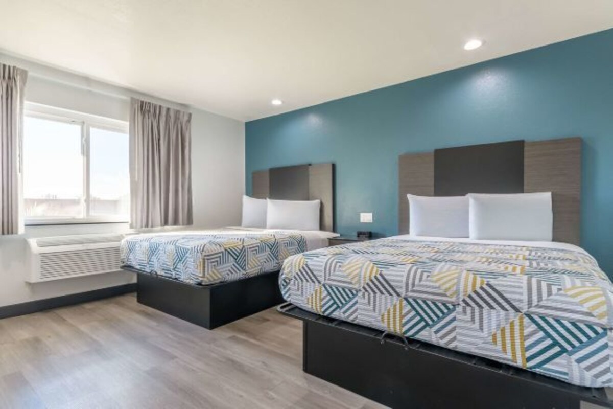 A hotel room with two beds, a window with curtains, a built-in air conditioner, and a blue accent wall.