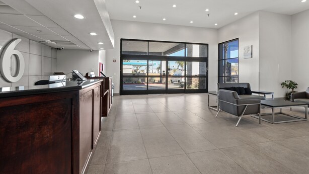 A modern reception area with a large number '6' sign, a reception desk, and a seating area with a view of the parking lot. - Studio 6 Suites Perris, CA (Perris)