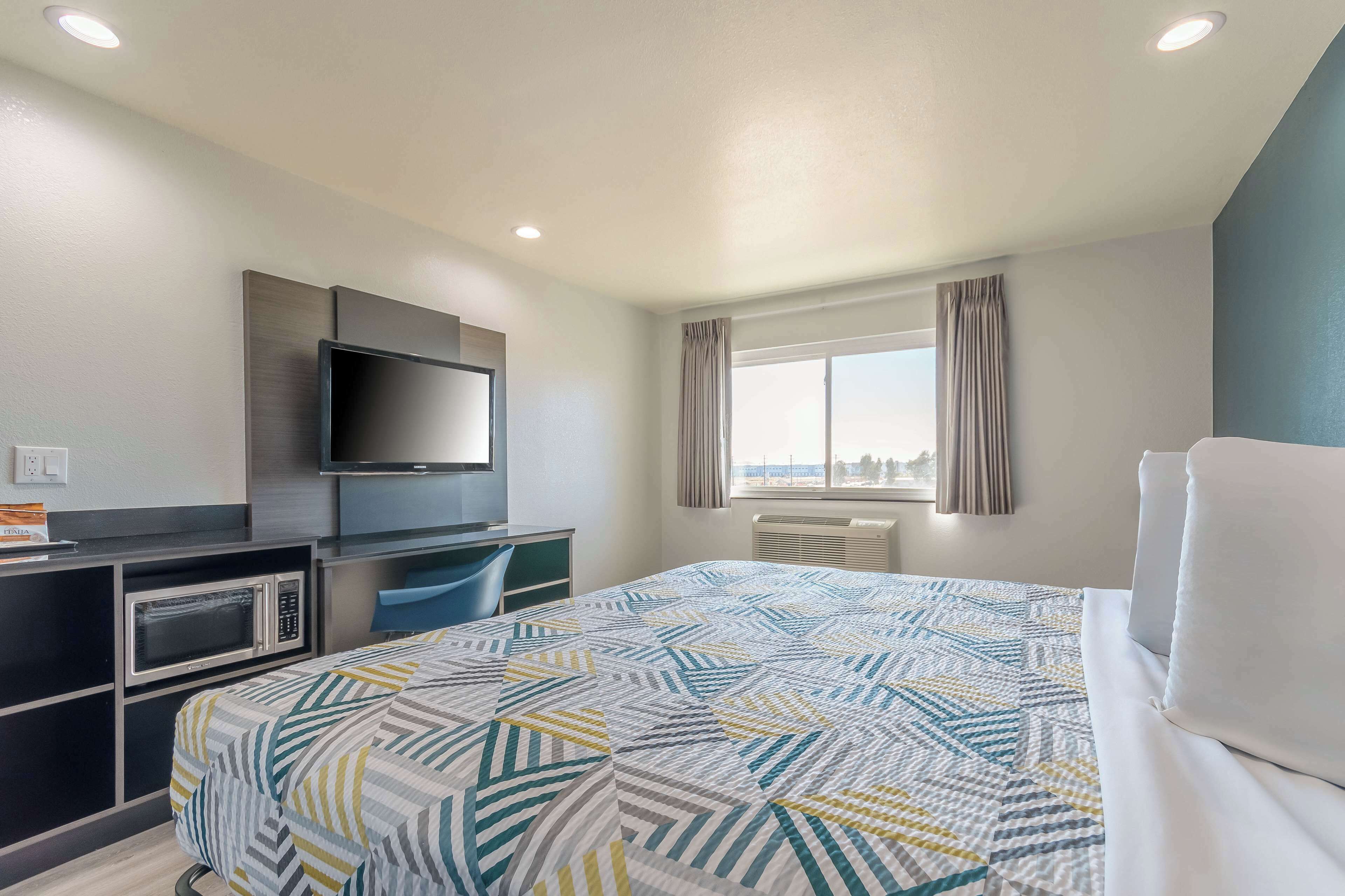 A hotel room with a bed, a TV mounted on the wall, a desk with a microwave, and a window with curtains.