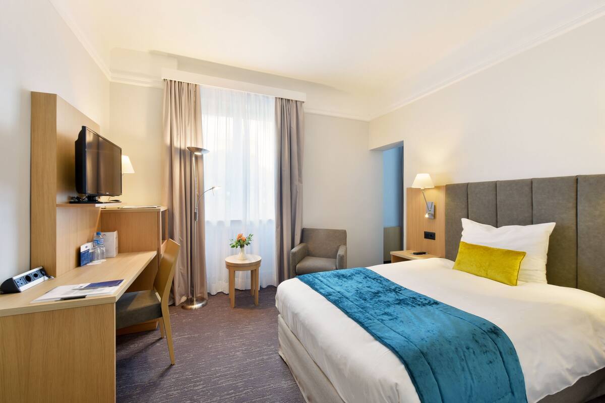 Ein Hotelzimmer mit Bett, Schreibtisch, Stuhl, Fernseher, Lampe und einem kleinen Tisch mit einer Blume.