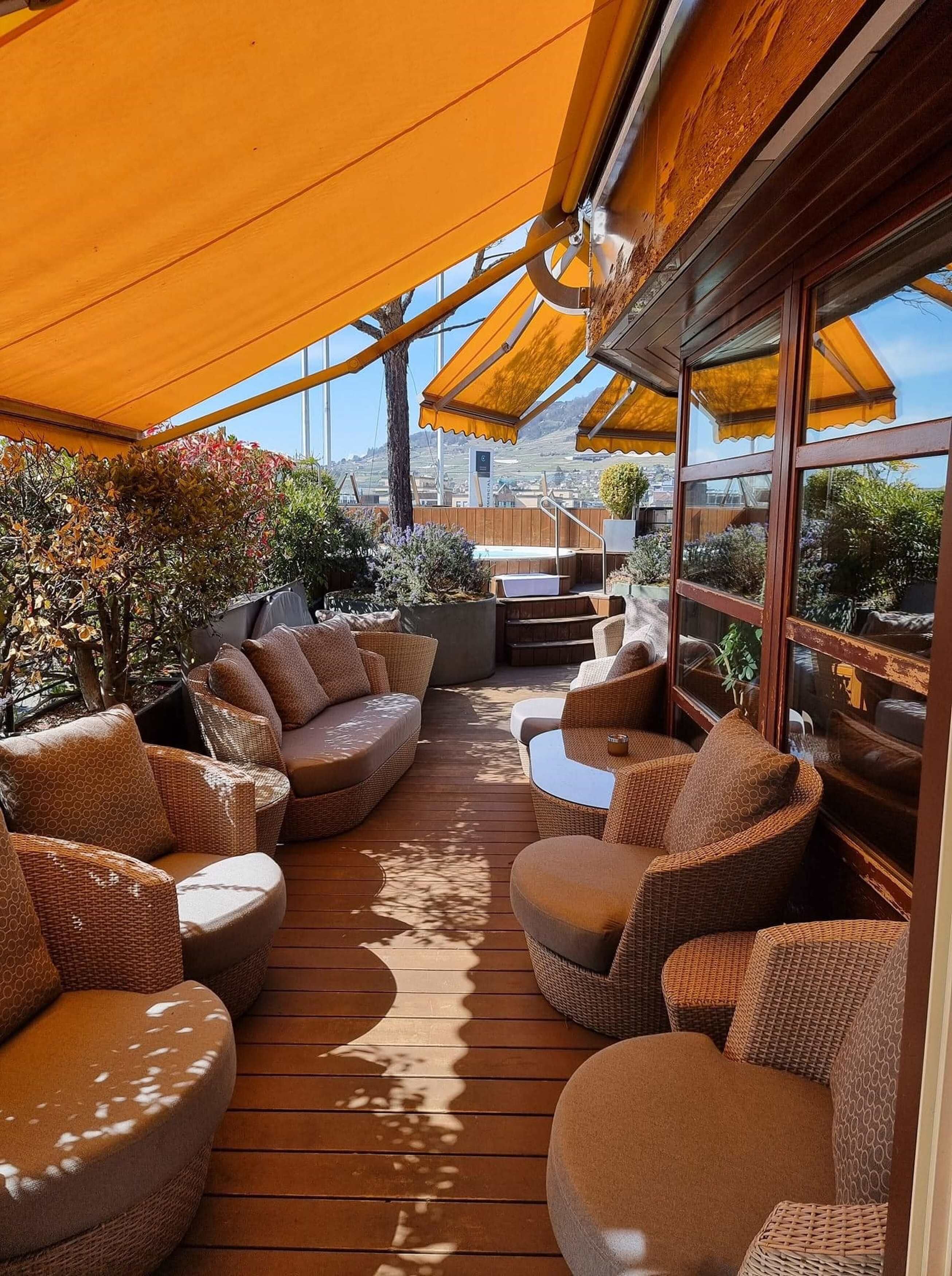 Terraza exterior con mobiliario de mimbre y suelo de madera, rodeada de toldos amarillos y plantas.